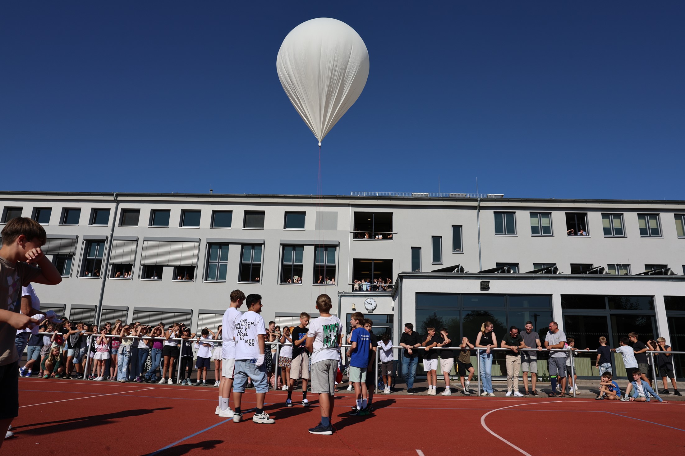 Eine Gruppe von Menschen steht auf einem roten Sportplatz, während ein großer weißer Ballon in den blauen Himmel aufsteigt.