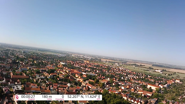 Luftaufnahme einer Stadtlandschaft mit zahlreichen roten Dächern und weitläufigen Feldern im Hintergrund bei klarem Himmel