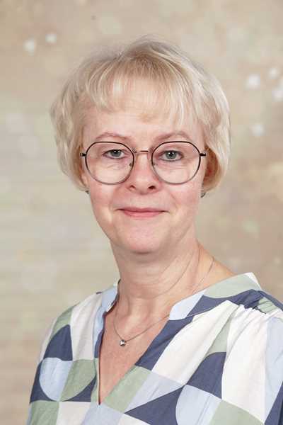 Eine lächelnde Frau mit Brille und blonden Haaren trägt ein gemustertes Hemd.