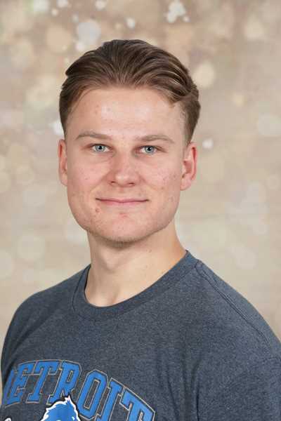 Ein junger Mann mit kurzen Haaren lächelt leicht, trägt ein graues Shirt.