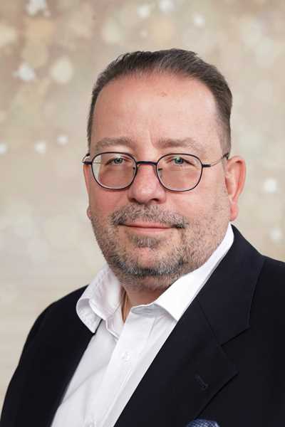 Ein Herr mit Brille und Bart lächelt, trägt ein dunkelblaues Sakko vor hellem Hintergrund