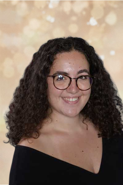 Eine lächelnde Frau mit lockigem Haar und Brille vor einem unscharfen Hintergrund.