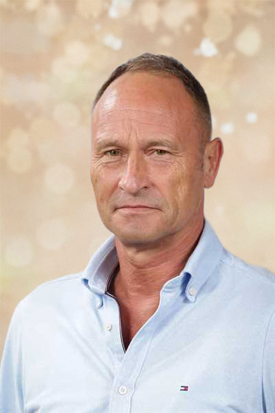 Ein Mann mit ernstem Gesichtsausdruck trägt ein hellblaues Hemd vor hellbraunem Hintergrund
