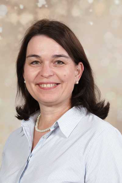Eine lächelnde Frau mit dunklem Haar, weißer Bluse und Perlenkette vor unscharfem Hintergrund.