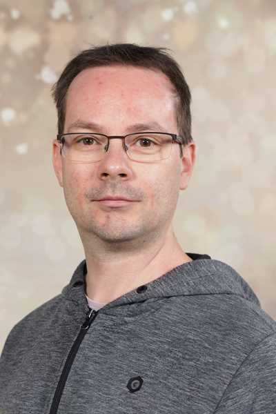 Ein Mann mit Brille lächelt leicht vor einem verschwommenen, beige Hintergrund.