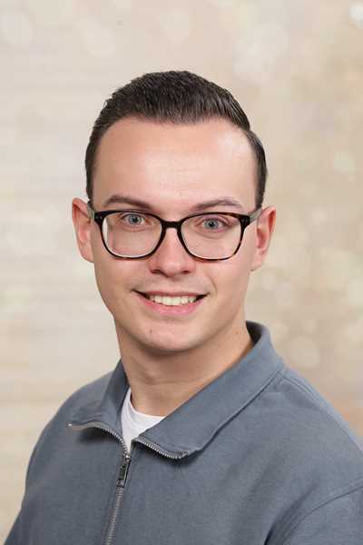 Ein junger Mann mit Brille lächelt freundlich, trägt ein graues Oberteil.