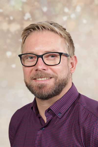 Ein Mann mit Brille, freundliches Lächeln, kariertes Hemd, hellbrauner Hintergrund
