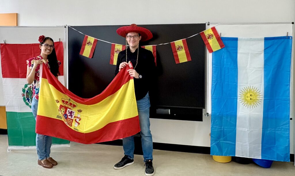 Zwei Personen lächeln, halten eine spanische Flagge, umgeben von Dekoration.