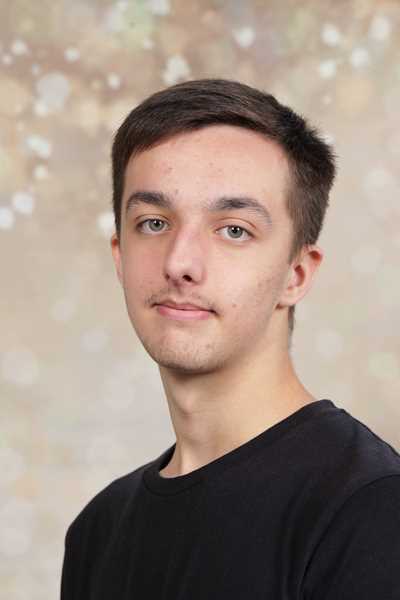 Ein junger Mann mit kurzem, dunklem Haar und neutralem Ausdruck trägt ein schwarzes T-Shirt vor einem verschwommenen Hintergrund.