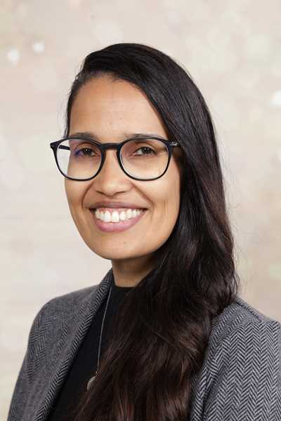 Eine Frau mit Brille lächeln, trägt einen grauen Blazer und hat dunkle Haare.
