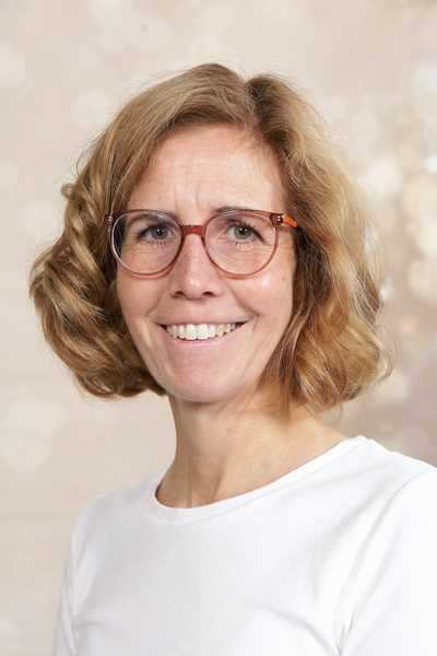 Eine lächelnde Frau mit lockigem, blondem Haar und Brille trägt ein weißes Oberteil.