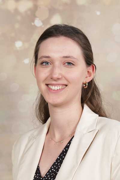 Eine lächelnde Person trägt einen cremefarbenen Blazer vor unscharfem Hintergrund.