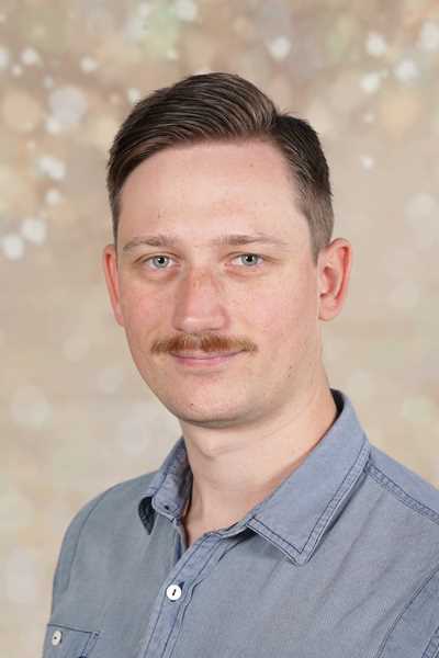 Ein Mann mit kurzem Haar und Schnurrbart lächelt sanft vor unscharfem Hintergrund