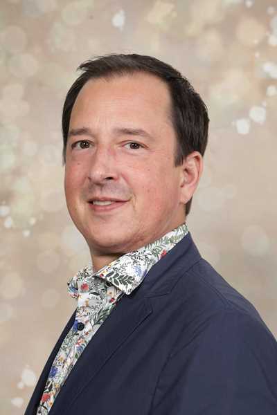 Ein lächelnder Mann in einem dunklen Anzug mit floralem Hemd vor unscharfem Hintergrund
