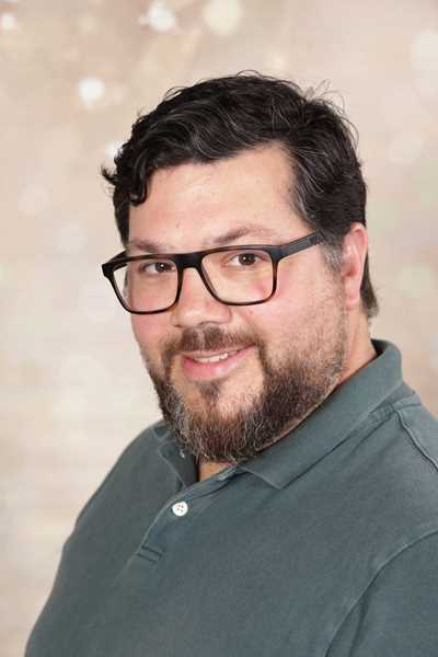 Ein Mann mit Brille und Bart lächelt in grauem Hemd vor hellem Hintergrund.