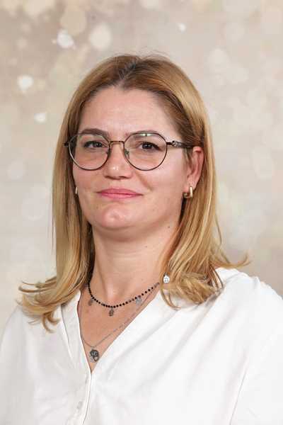 Eine lächelnde Frau mit blonden Haaren und Brille trägt eine weiße Bluse vor einem bokeh Hintergrund