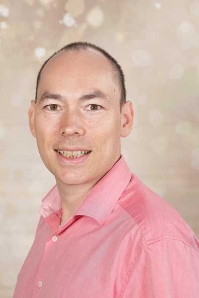Eine lächelnde Person in einem rosa Hemd, vor einem verschwommenen Hintergrund.