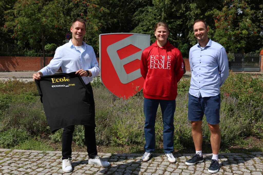 Drei Männer stehen lächelnd vor einem Schild in einem Garten.