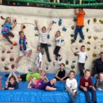 Eine Gruppe fröhlicher Kinder klettert auf einer bunten Kletterwand in einer Turnhalle.