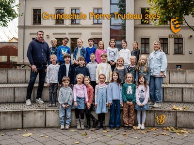 Eine fröhliche Kindergruppe mit zwei Erwachsenen steht vor einer Schule, viele lächeln