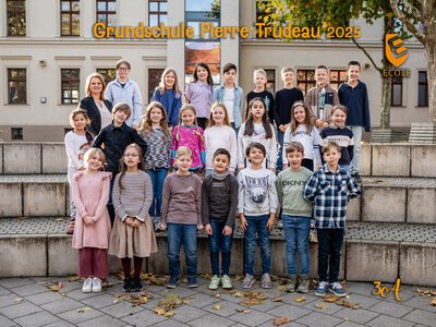 Eine fröhliche Grundschulklasse mit Lehrerin posiert vor einem Schulgebäude in Herbstfarben