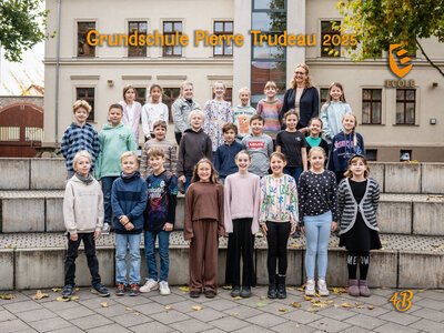 Eine Gruppe lächelnder Grundschulkinder und eine Frau stehen vor einer Schule auf Treppen