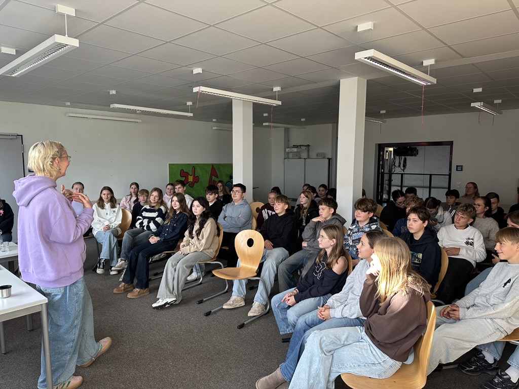 Eine ältere Frau mit blondem Haar spricht lächelnd vor einer Gruppe Jugendlicher