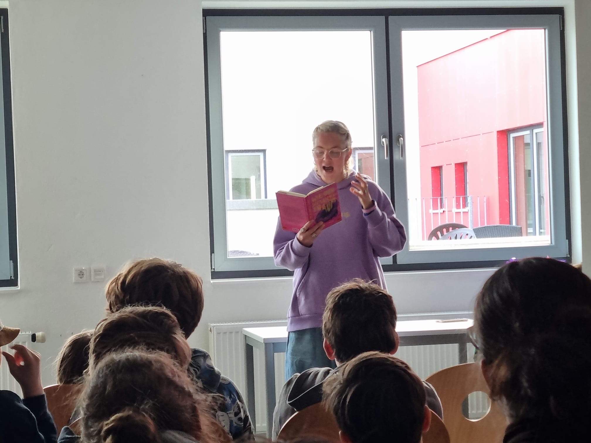 Eine Person mit Brille liest lächelnd aus einem pinkfarbenen Buch vor einer Schulklasse vor