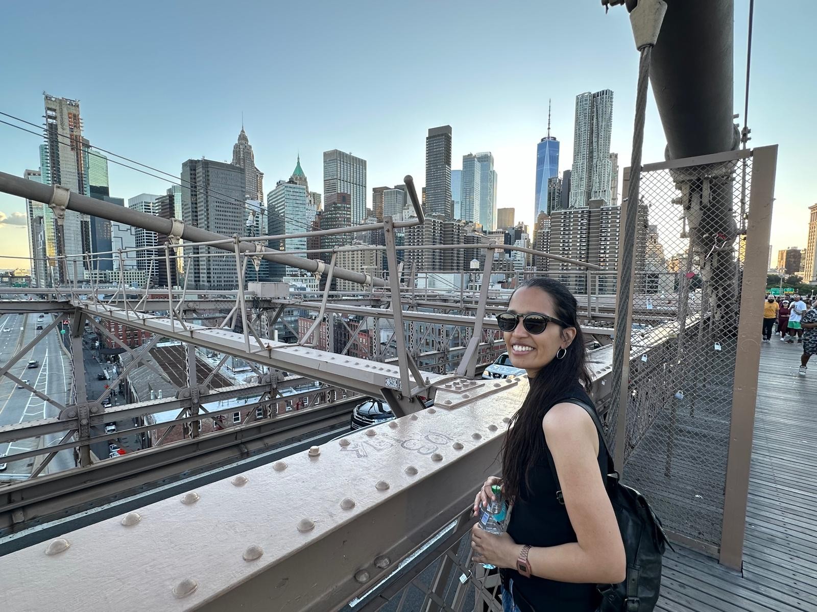 Eine Frau mit langen dunklen Haaren und Sonnenbrille lächelt auf der Brooklyn Bridge vor der Skyline von New York und hält eine Wasserflasche in der Hand