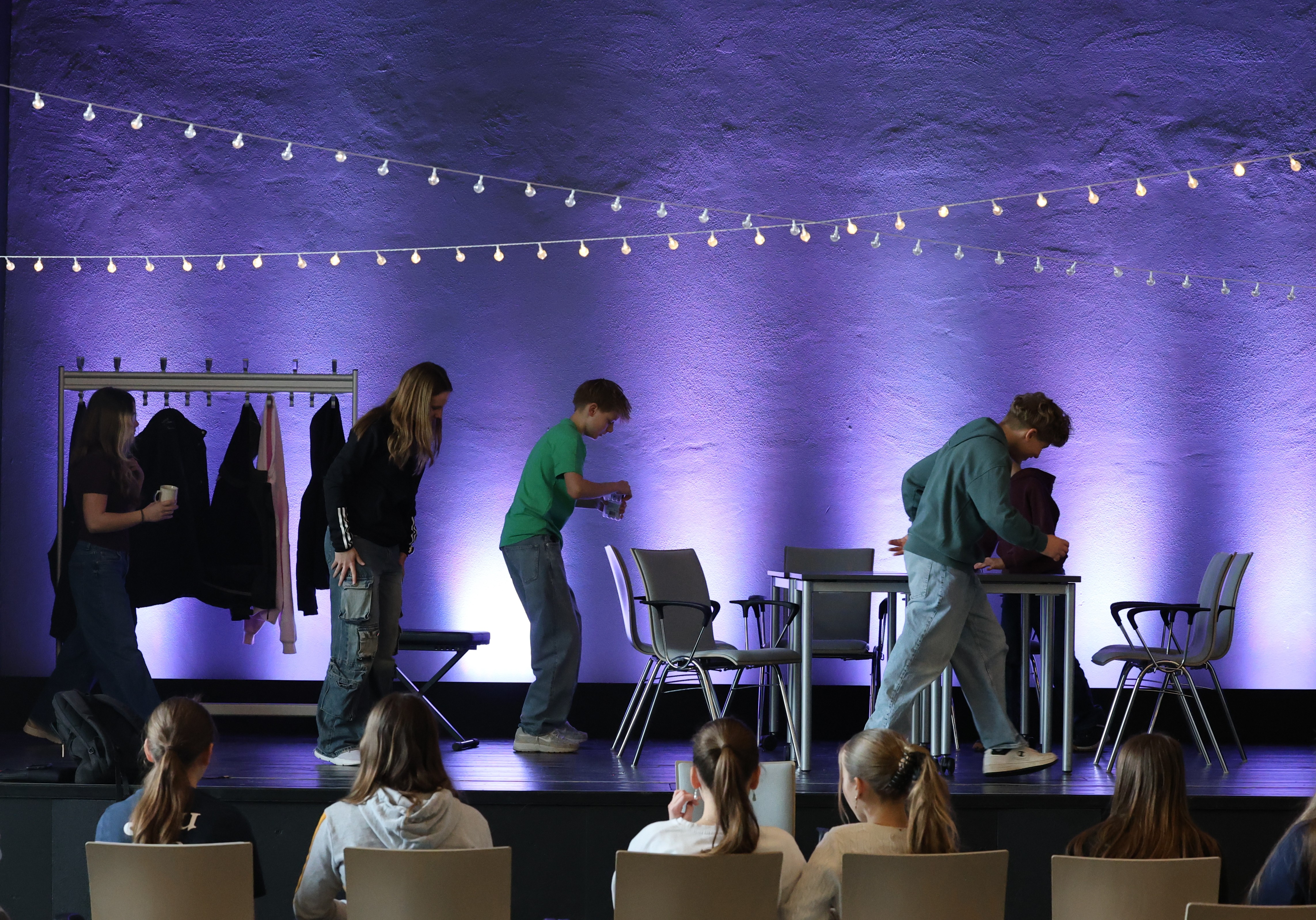 Fuenf junge Menschen auf einer Buehne mit lila Beleuchtung und Lichterketten wirken konzentriert und fuehren scheinbar eine Theaterszene auf