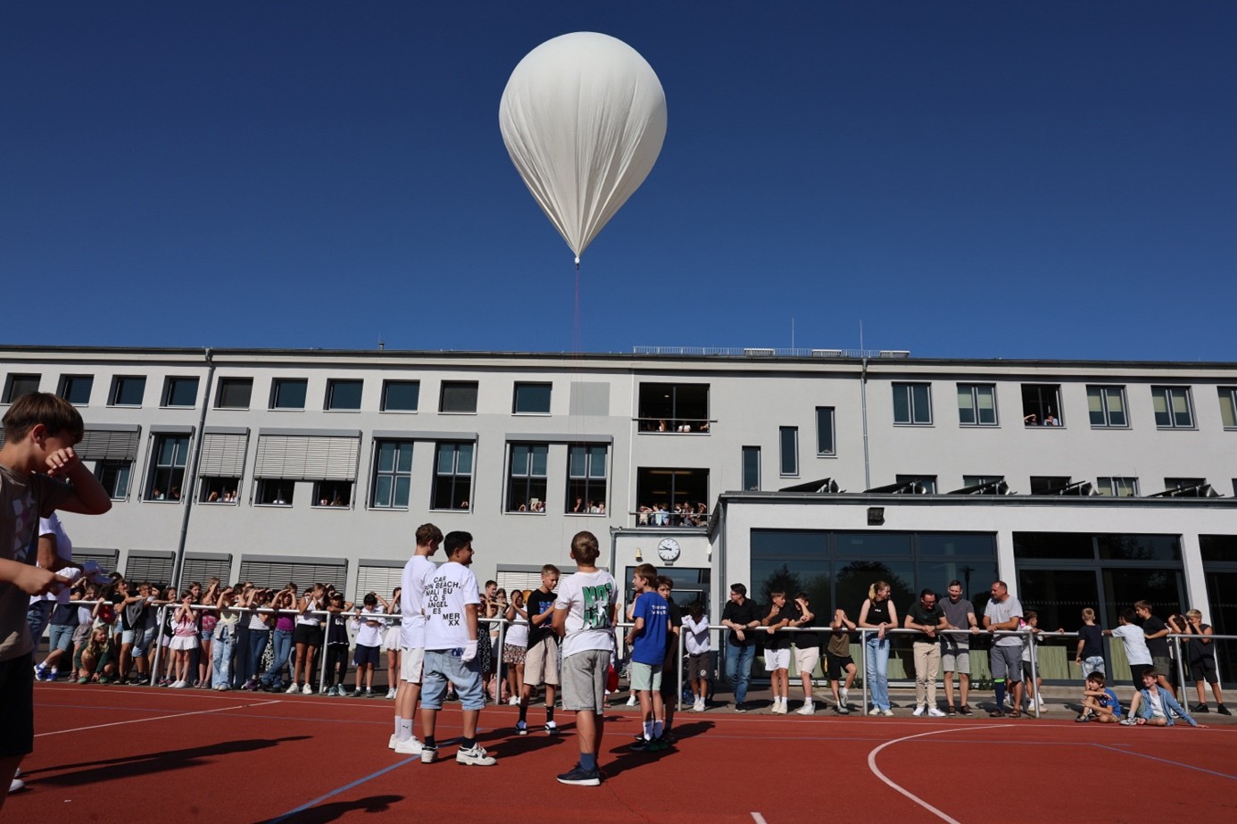 Mehrere Kinder stehen auf einem roten Sportplatz vor einer Schule, über ihnen schwebt ein weißer Wetterballon am klaren blauen Himmel
