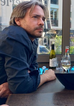 Ein nachdenklicher Mann mit Bart und grauem Hemd sitzt an einem Tisch mit Getränken