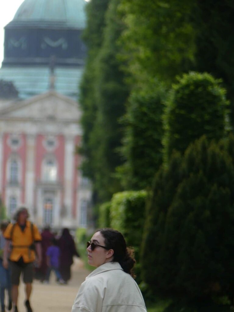 Eine Frau mit dunklen Haaren und Sonnenbrille blickt entspannt vor einem Schloss