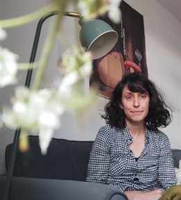 Frau mit lockigen dunklen Haaren sitzt ruhig auf einem grauen Sofa neben einer Lampe