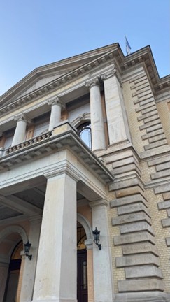 Das Bild zeigt ein historisches Gebäude mit hohen Säulen und heller Fassade vor blauem Himmel