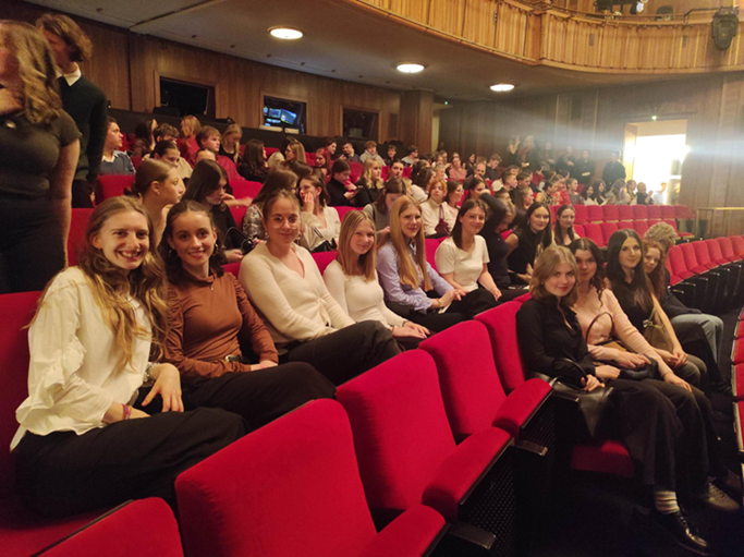 Eine Gruppe junger Frauen lächelt freundlich auf roten Kinosesseln im Theater