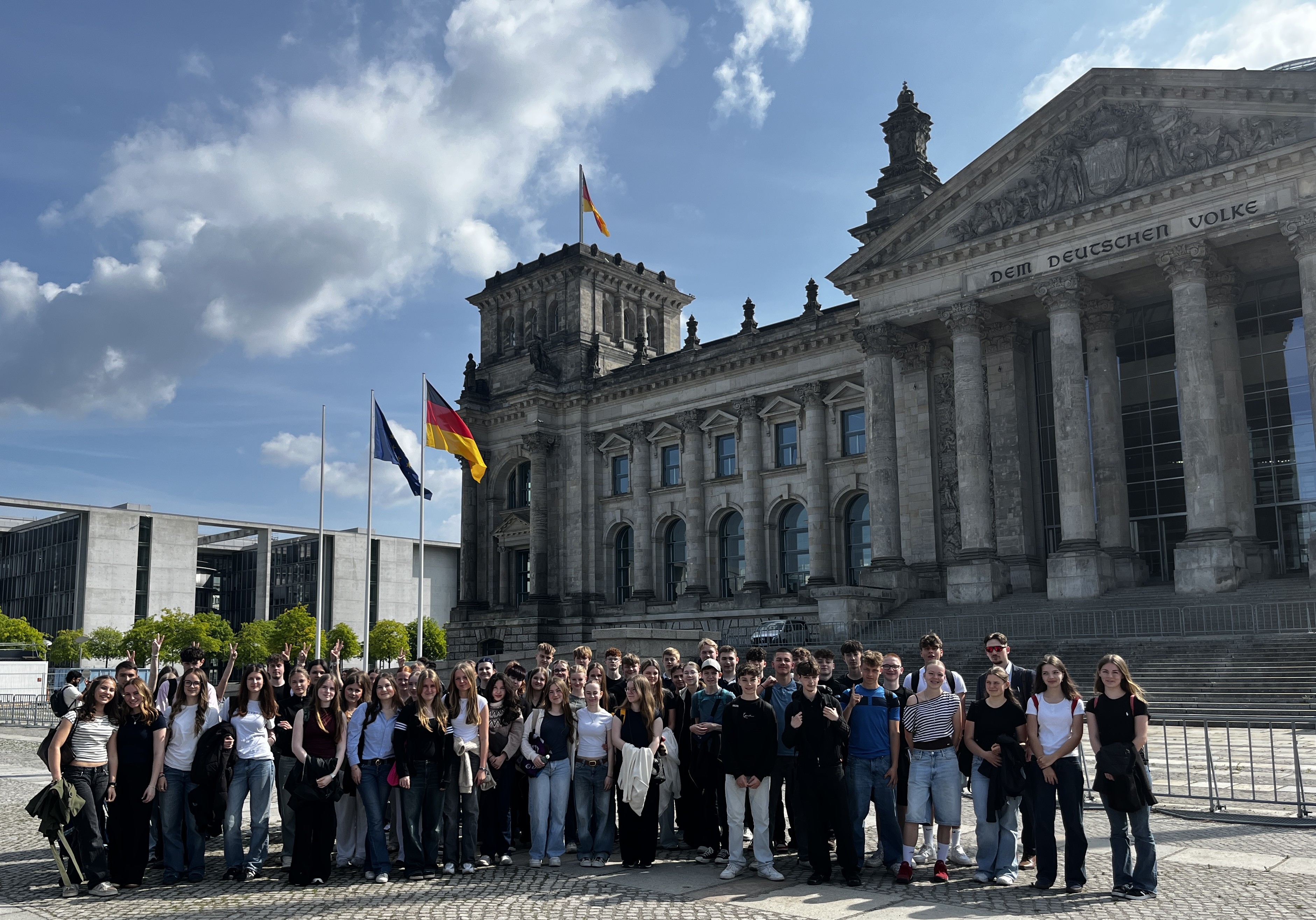 Lachende Menschengruppe vor grauem Reichstag mit Flaggen und blauem Himmel