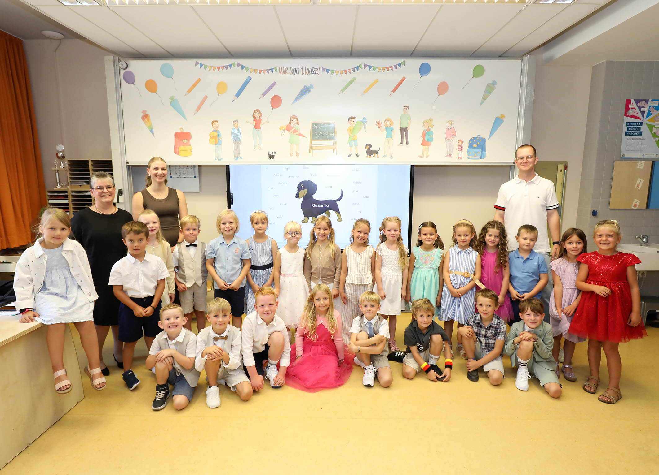 Klassenfoto der ersten Klasse mit festlich gekleideten Kindern und drei Lehrkraeften