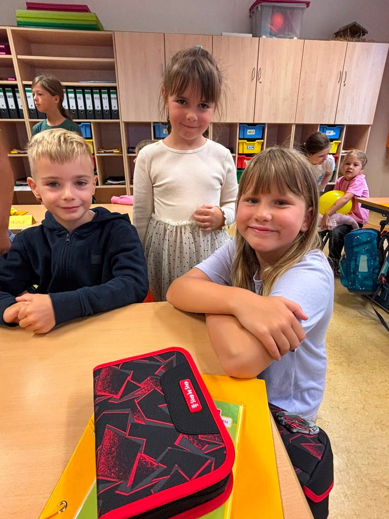 Drei laechelnde Kinder sitzen im Klassenzimmer an einem Tisch mit Federmaeppchen