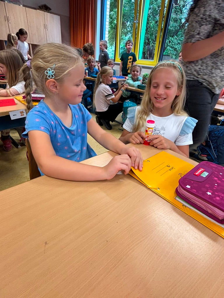 Zwei laechelnde blonde Maedchen sitzen im Klassenzimmer an einem Holztisch