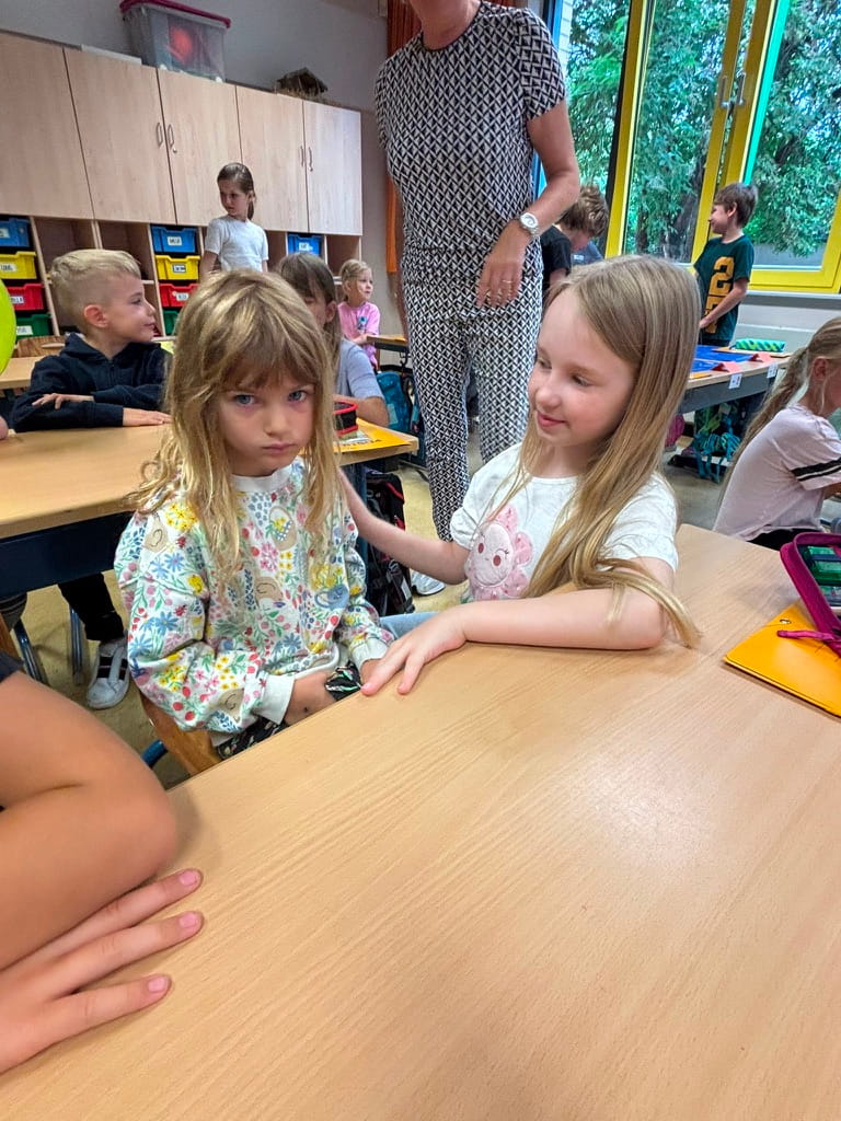 Zwei blonde Maedchen sitzen laechelnd am Tisch im hellen Klassenzimmer
