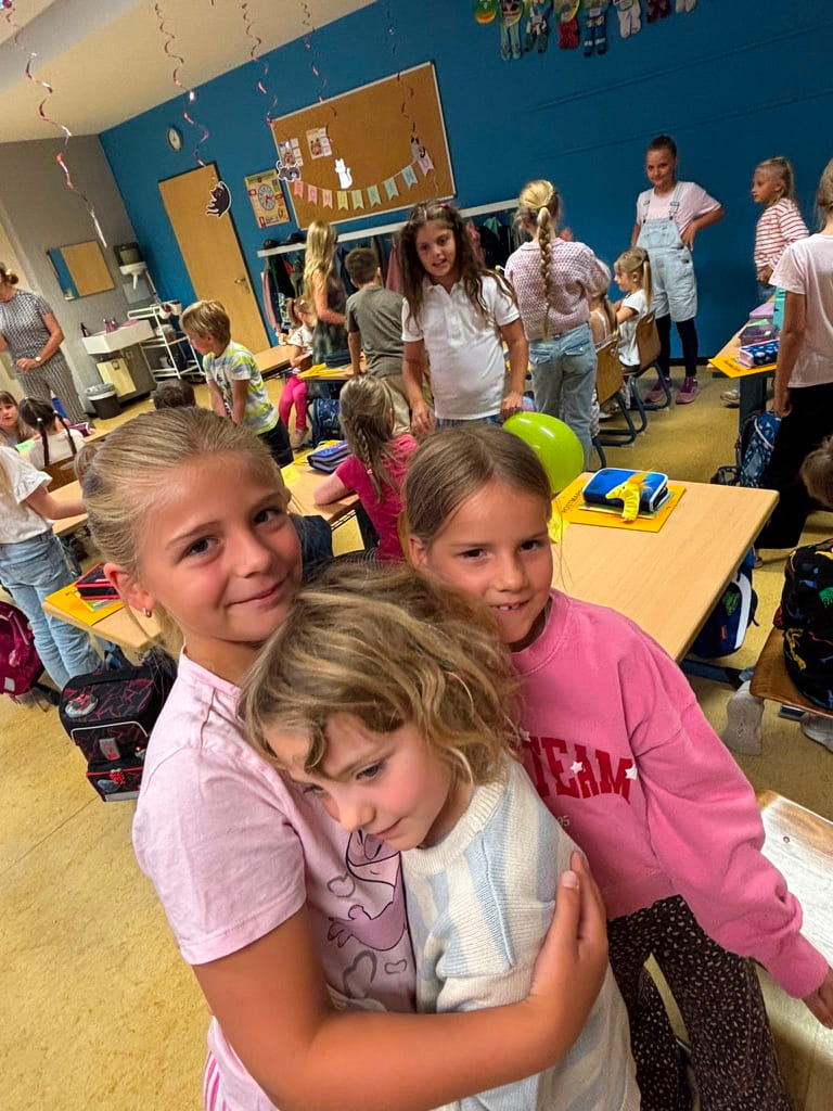 Drei laechelnde Kinder in rosa und weiss umarmen sich im Klassenzimmer