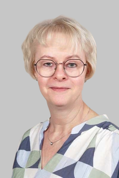 Eine laechelnde Frau mit blonden Haaren und Brille traegt eine gemusterte Bluse