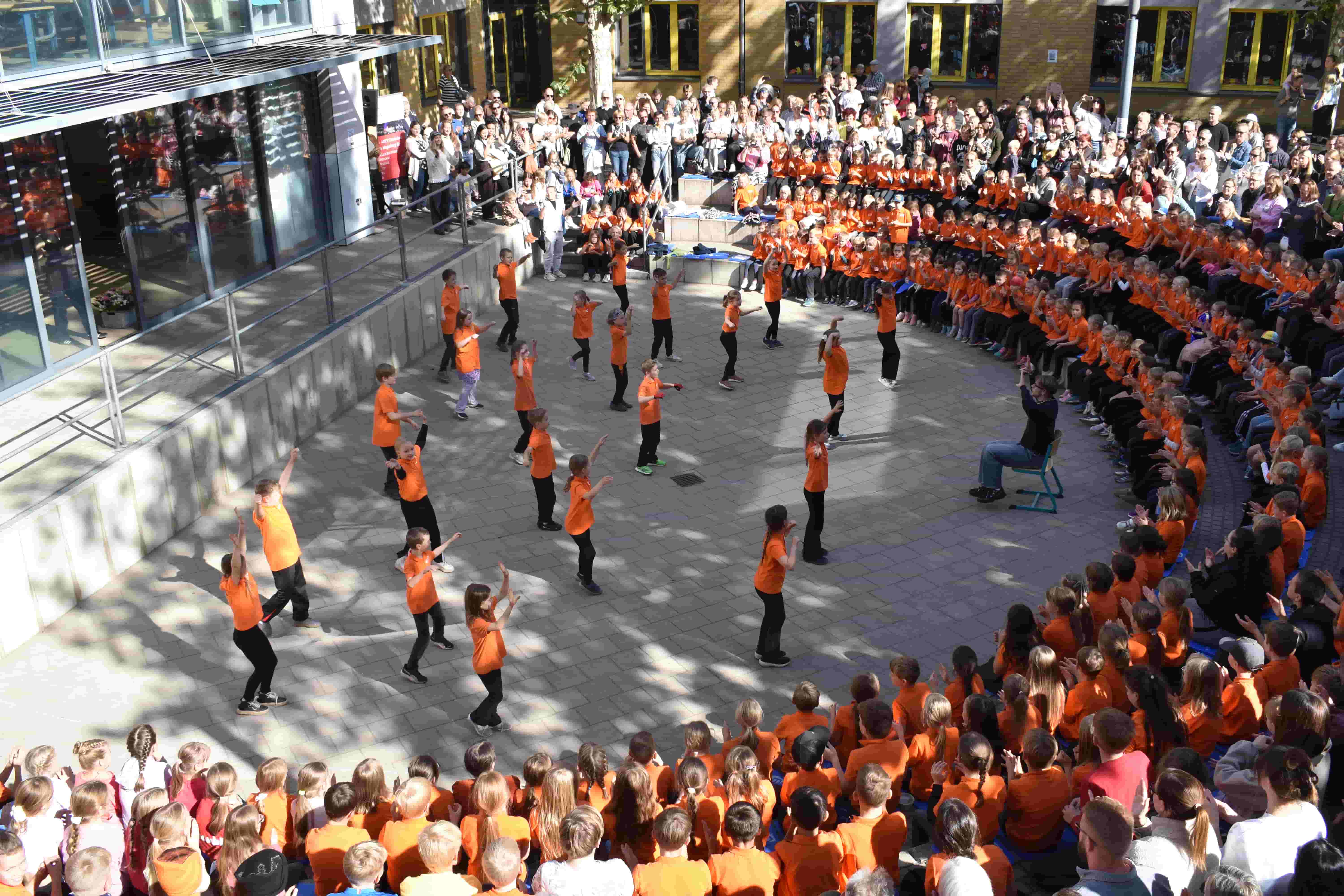 Kinder in orangefarbenen Shirts tanzen frohlich im Schulhof vor Publikum