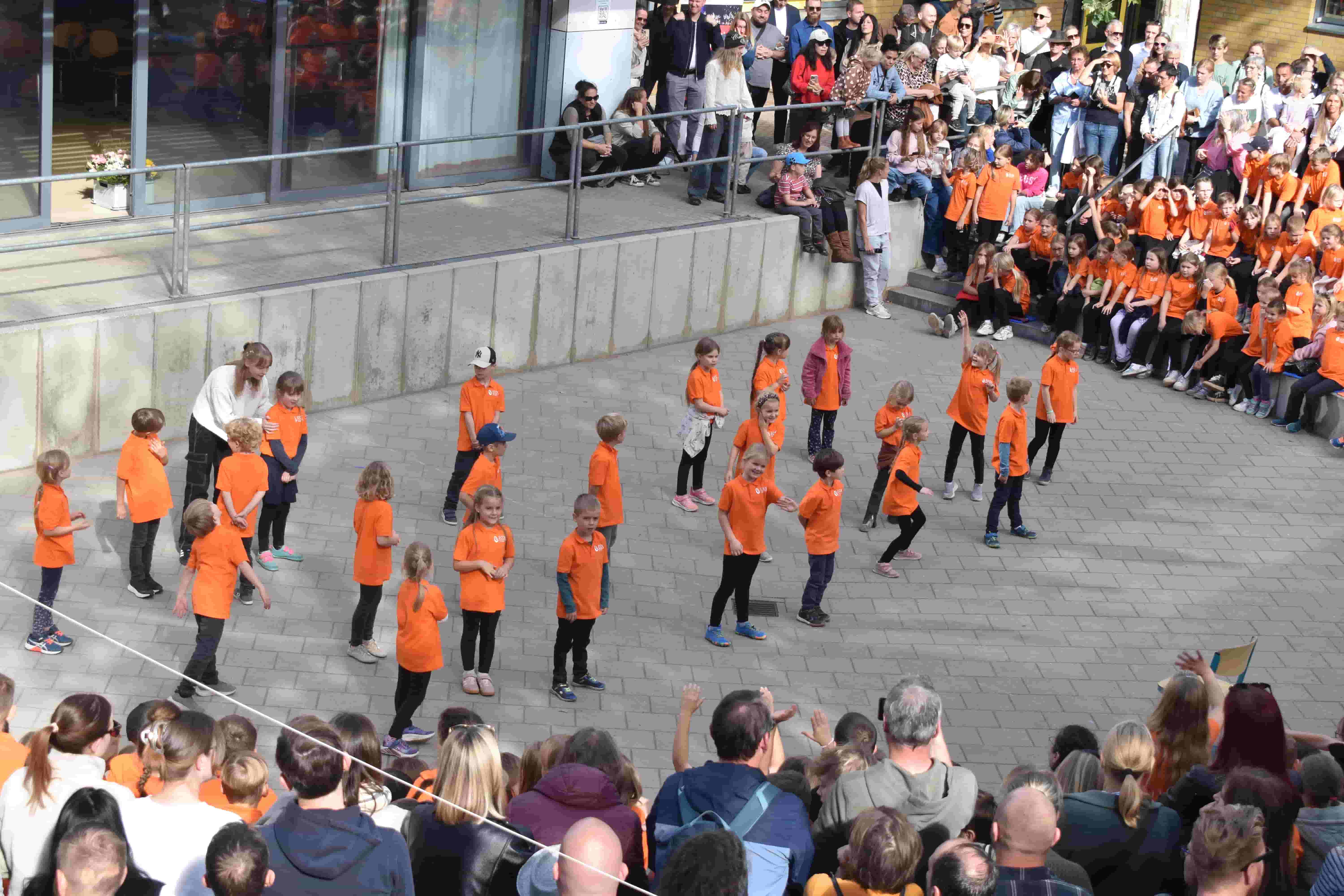 Kinder in orangefarbenen Shirts tanzen froehlich vor einem Publikum im Freien