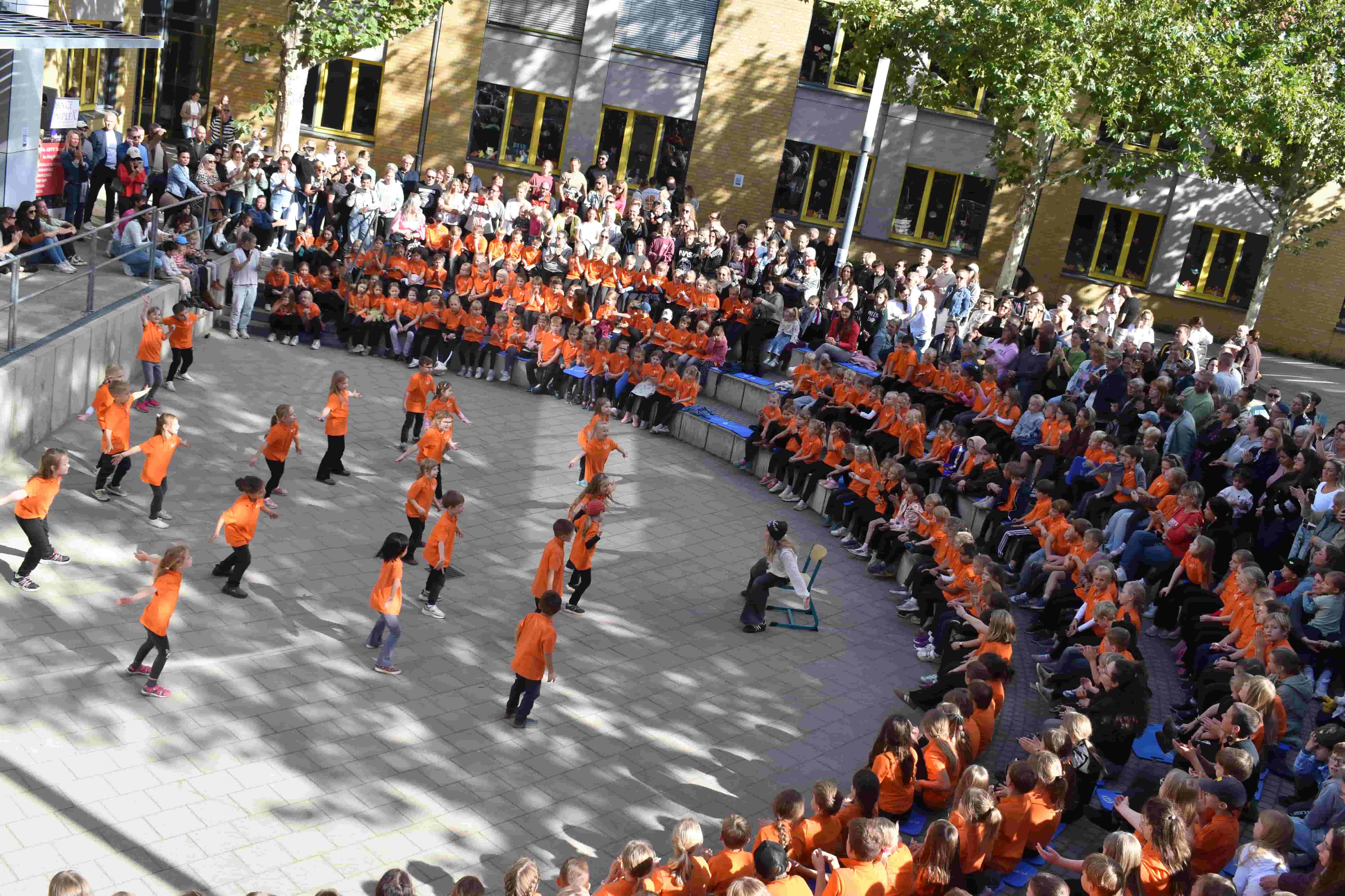 Kinder in orangefarbenen Shirts tanzen auf einem Schulhof vor Publikum