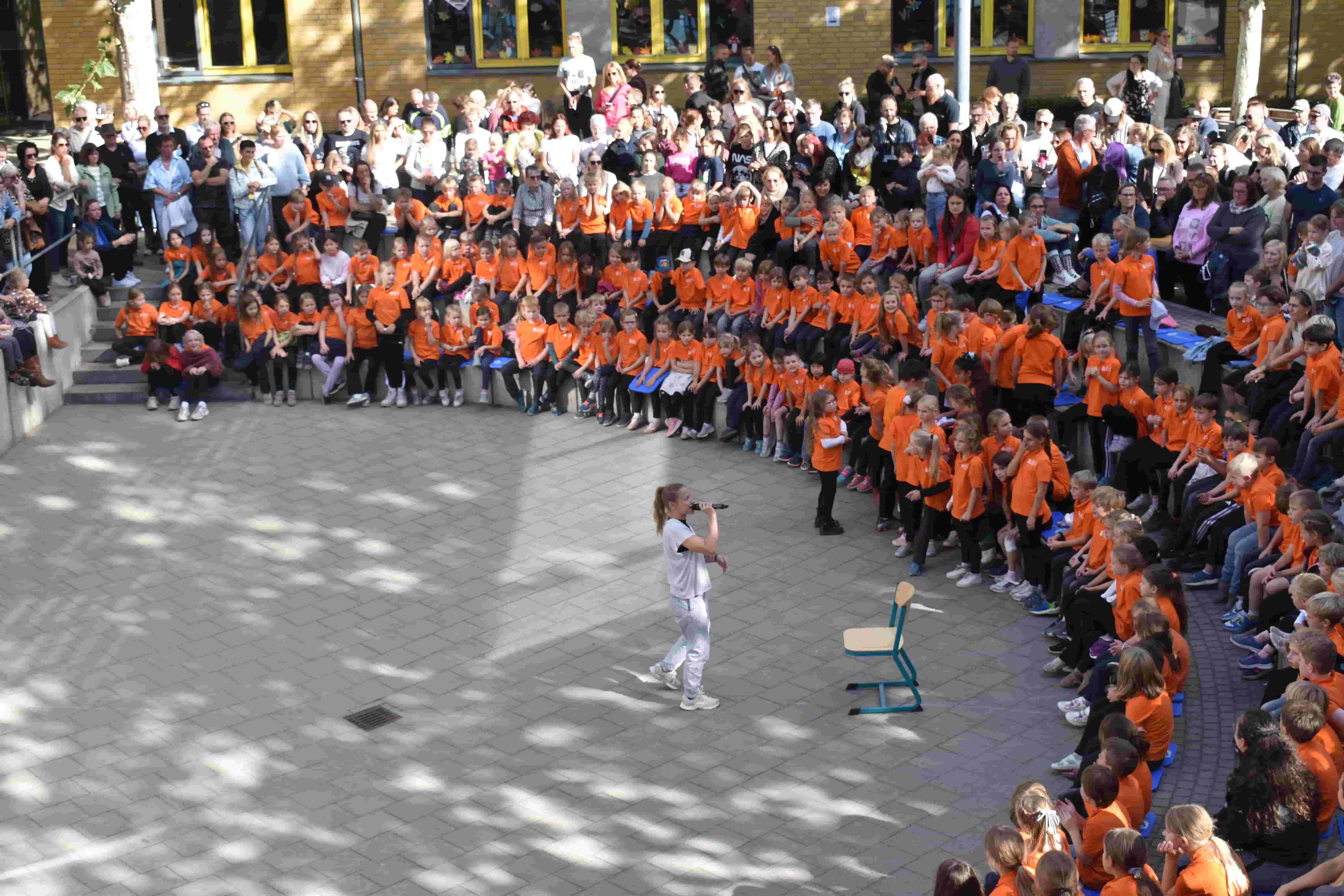 Frau in Weiss singt mit Mikrofon vor Kindern in orangefarbenen Shirts