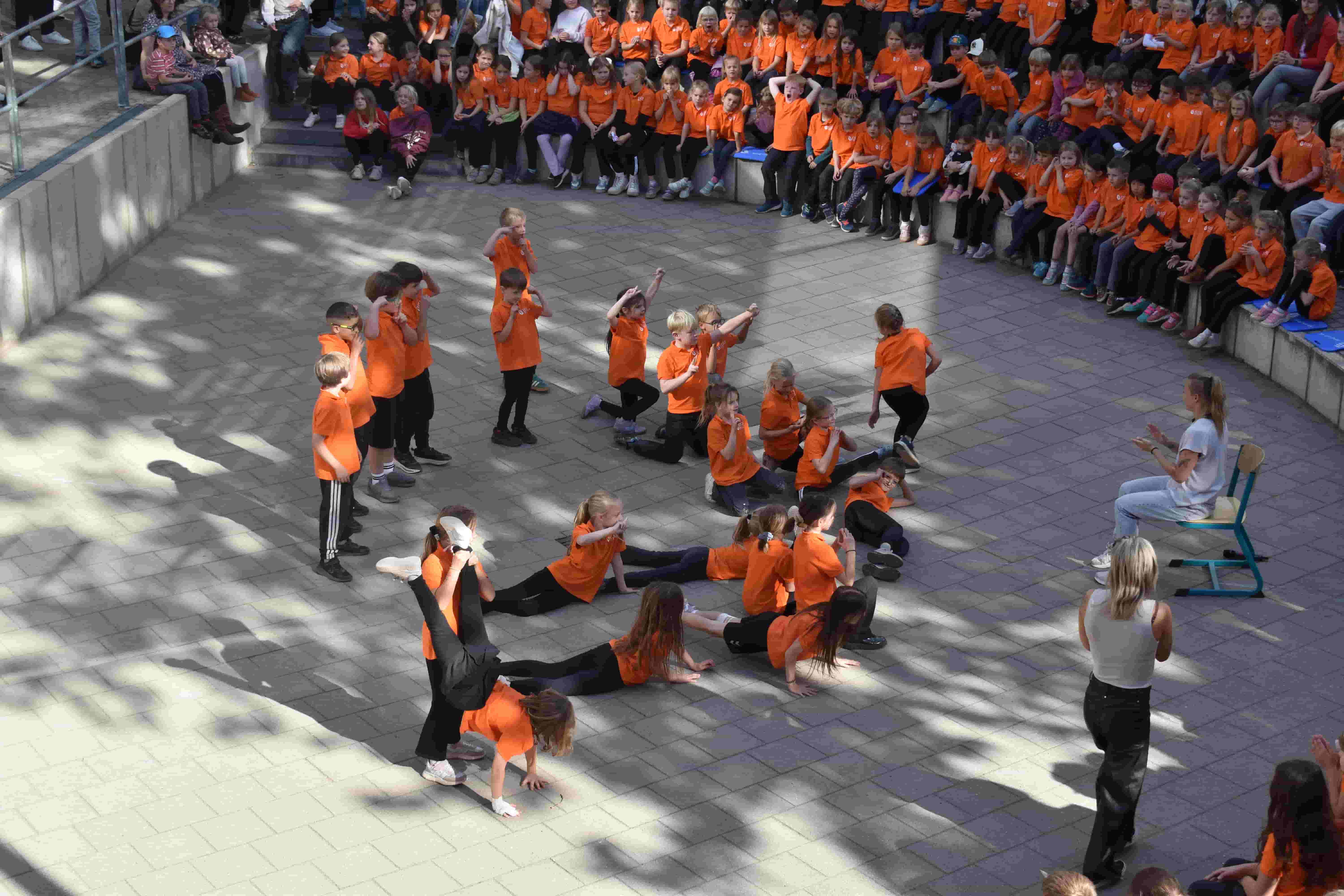 Kinder in orangefarbenen Shirts tanzen auf einem Schulhof vor Zuschauern