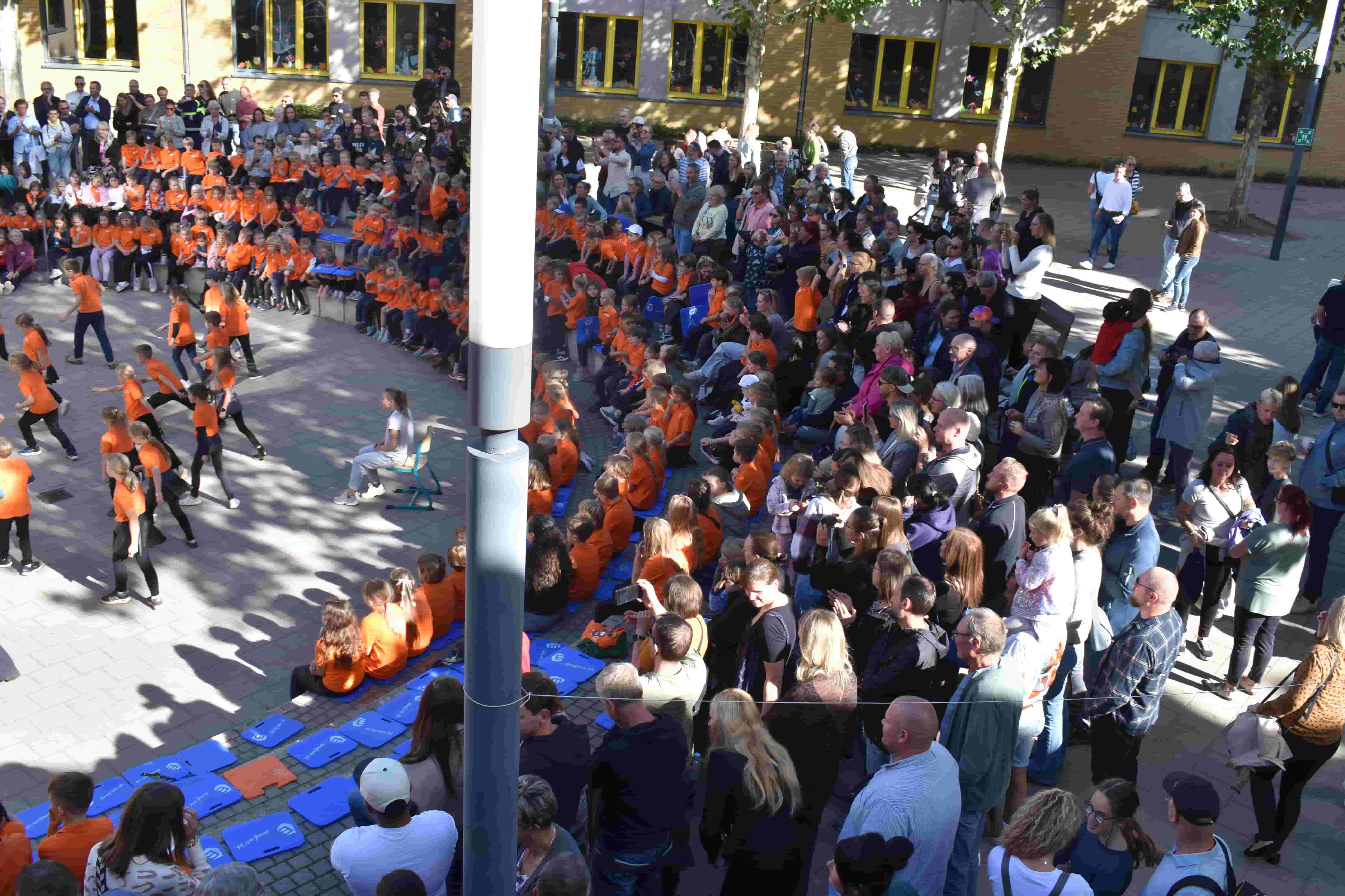 Kinder in orangefarbenen Shirts tanzen vor Zuschauern auf einem Schulhof