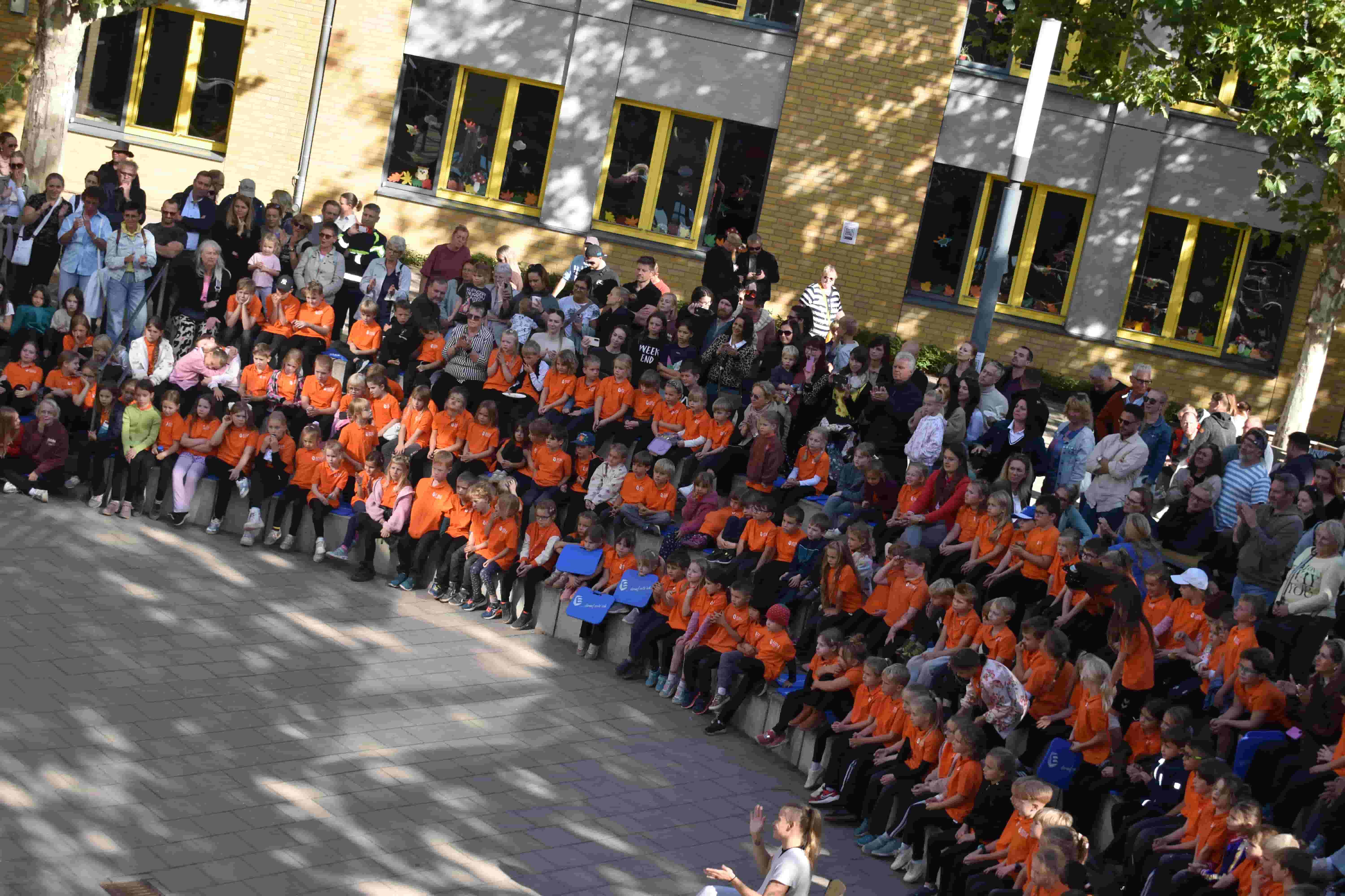 Viele Kinder in orangefarbenen Shirts sitzen mit Erwachsenen vor einem Schulgebaeude
